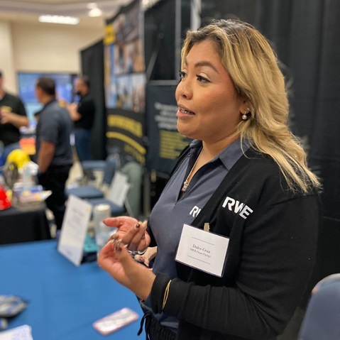 A woman in a dark RWE sweater stands at a booth, engaging with attendees at an event, with a name tag visible.