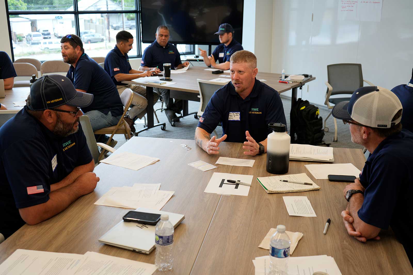 A group discusses around a table in a bright room filled with papers and drinks, showcasing teamwork and collaboration.