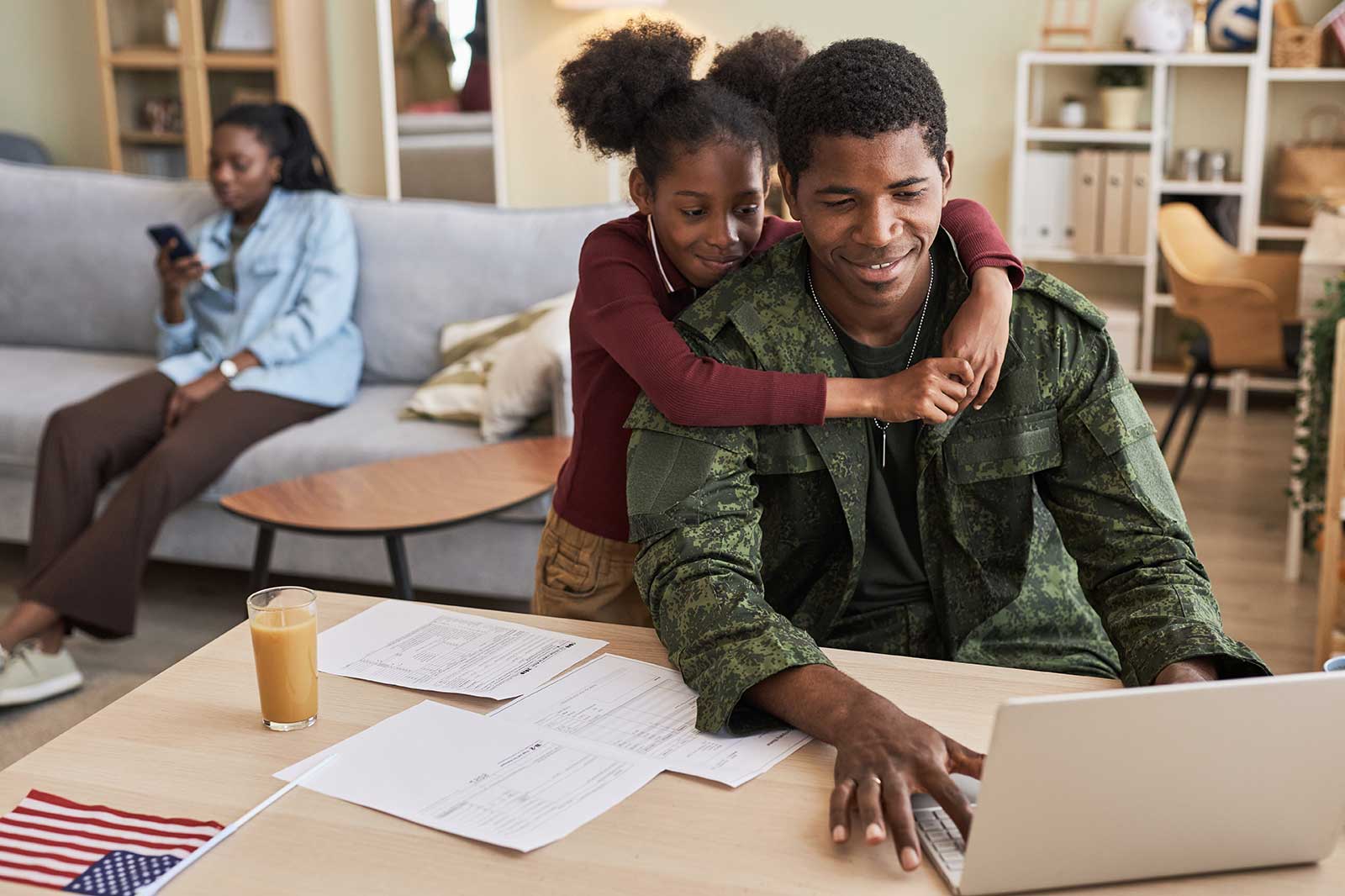 A man in military uniform works on a laptop at a desk while a girl hugs him. A woman sits on a couch using her phone.