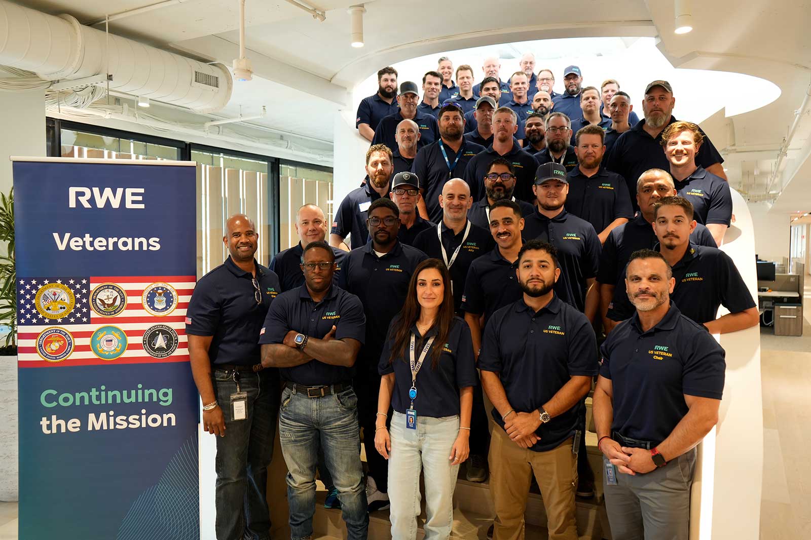 A group of veterans stands on a staircase, wearing navy blue polo shirts with RWE branding. A banner reads 'RWE Veterans'.