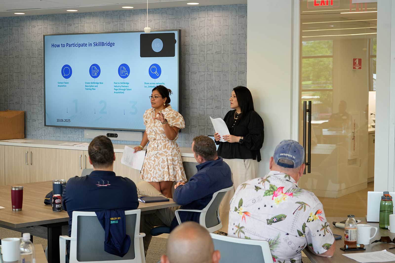 A presentation in a modern office with a screen showing instructions for participating in SkillBridge. Attendees are engaged.