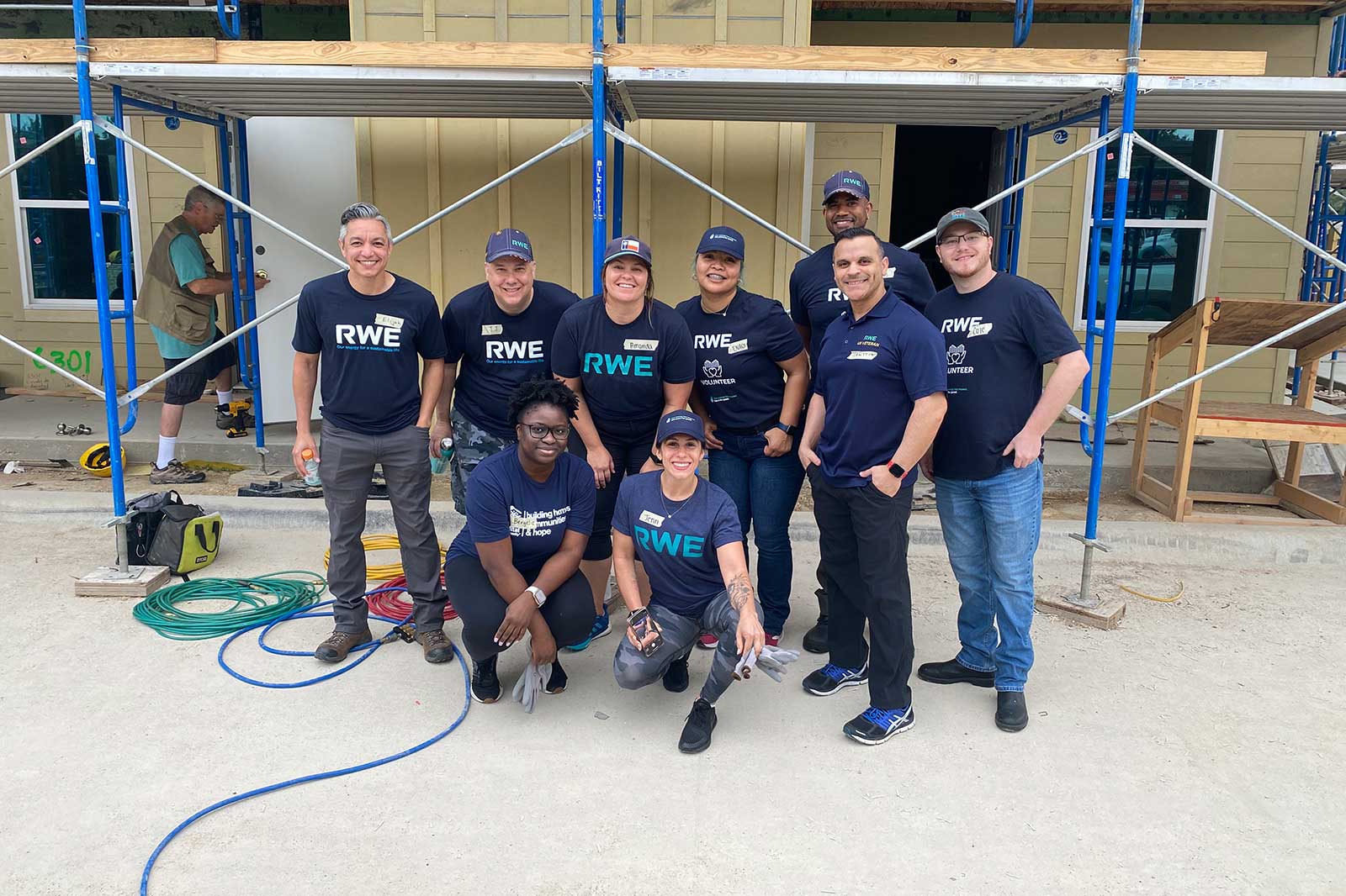 A group of volunteers wearing RWE shirts poses for a photo at a construction site with scaffolding in the background.