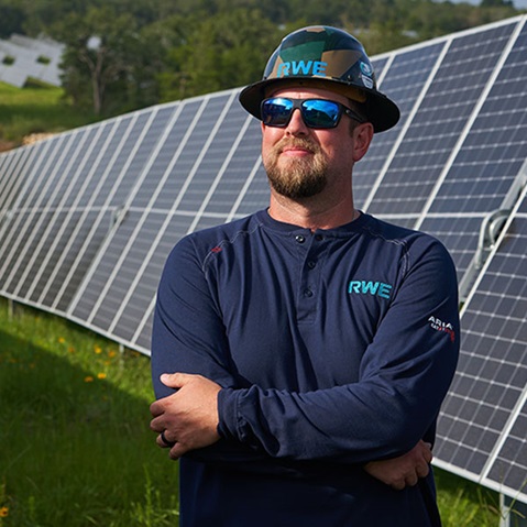 A technician stands confidently in front of a solar panel array, wearing a hard hat and a long-sleeve shirt with the RWE logo.