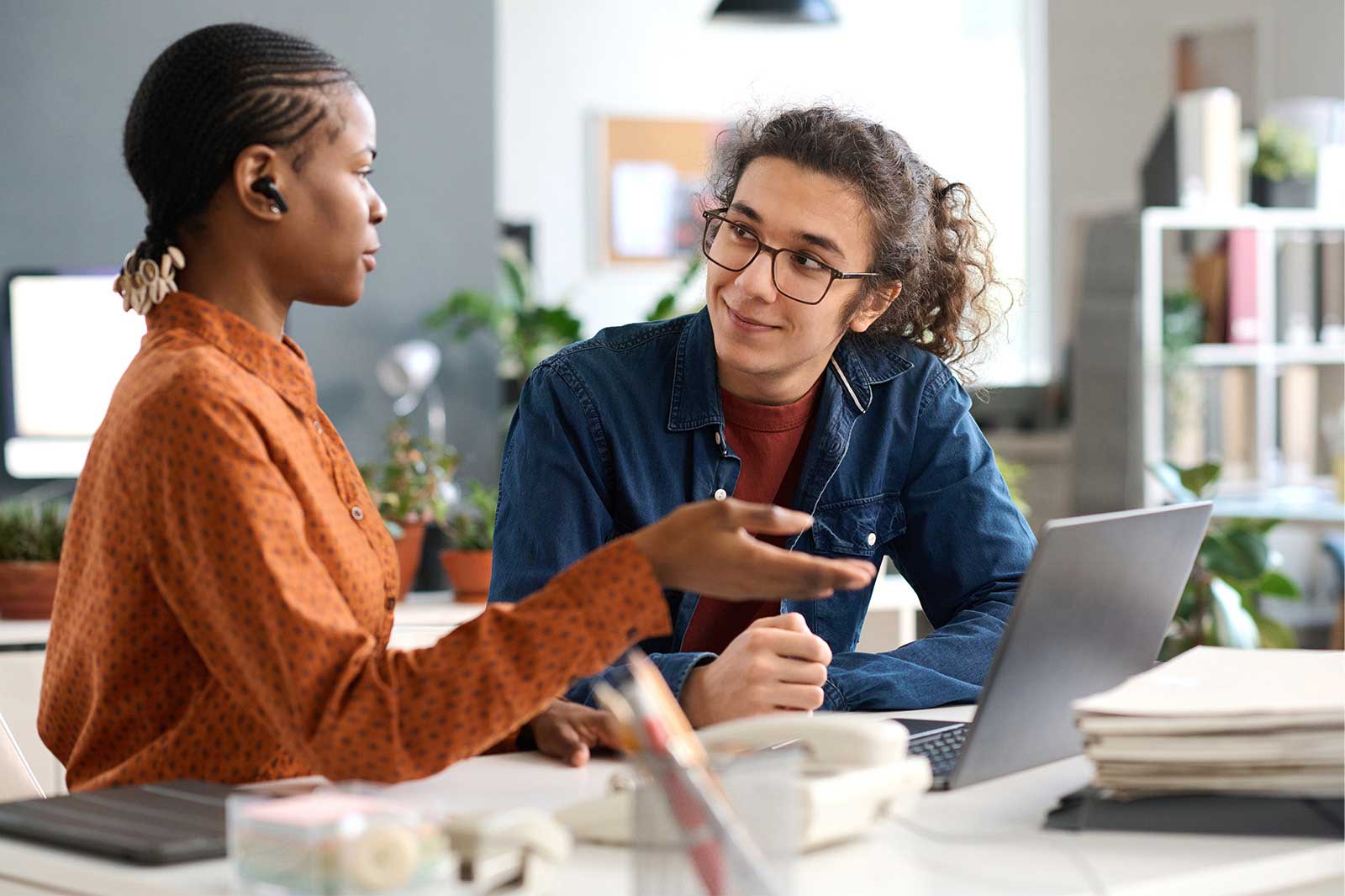 Two individuals are discussing and working on a laptop in a modern office, surrounded by plants and office supplies.
