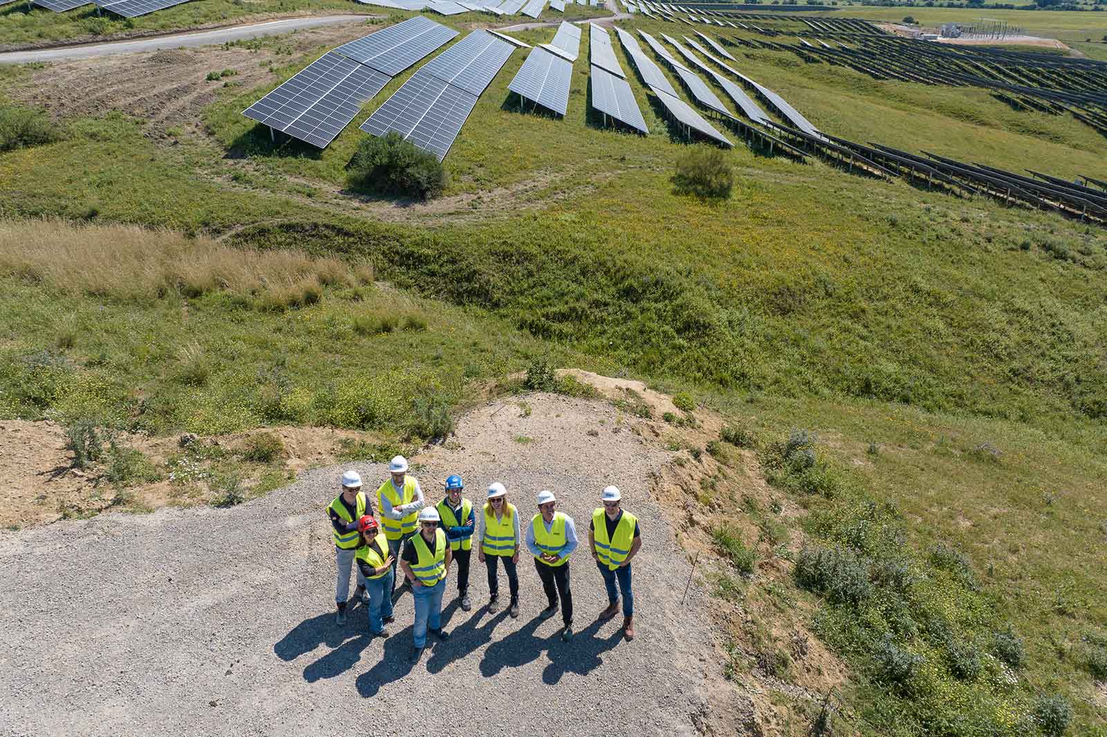 A group of seven individuals wearing safety vests and helmets stand on gravel, with solar panels in the background on grassy hills.