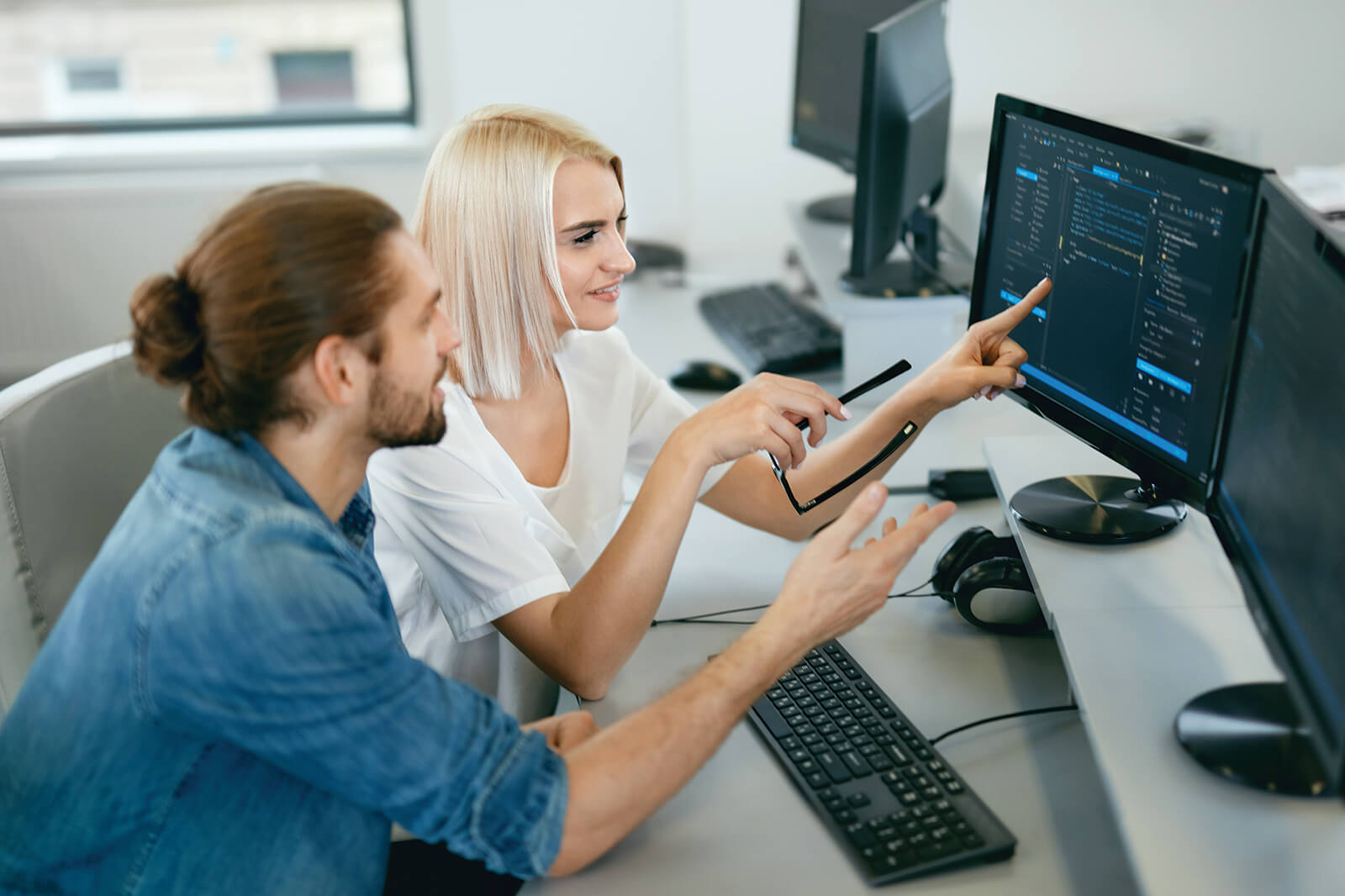 Two individuals collaborate at a computer, discussing content displayed on the screen filled with written code.