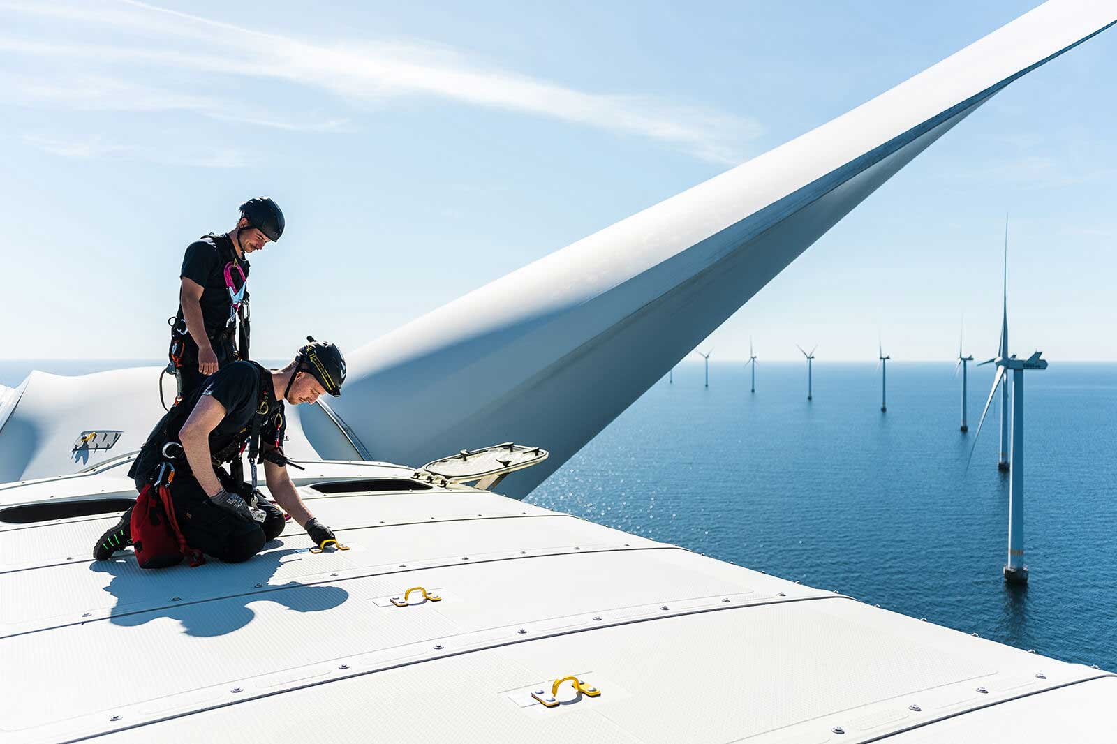 Two technicians work atop a wind turbine, overlooking multiple turbines in the ocean under a clear blue sky.