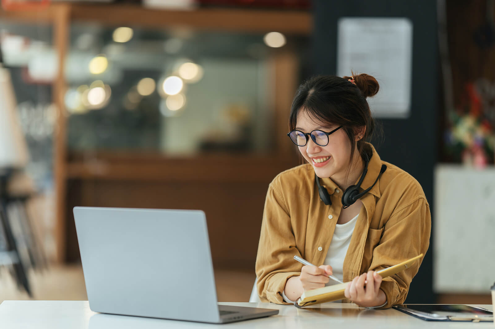 A person sits at a café table with a laptop and notepad, jotting down notes.